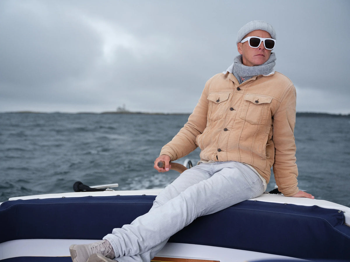 Woman wearing white VIDAR Ample sunglasses, a beige jacket, grey knit beanie and scarf, sitting on the edge of a boat beneath an overcast sky.